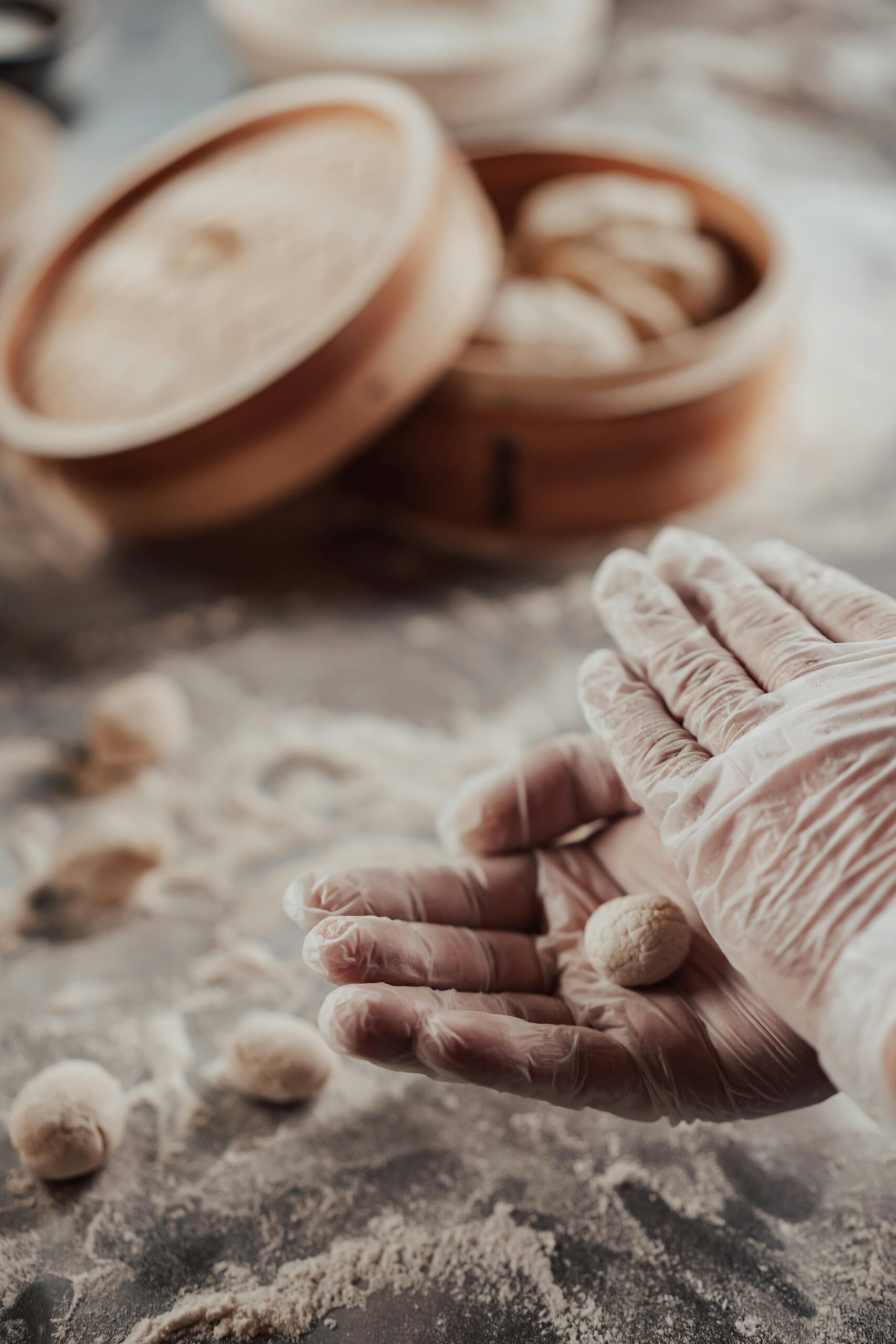 Gloved hands shaping dough for dumplings near a bamboo steamer in a kitchen setting.